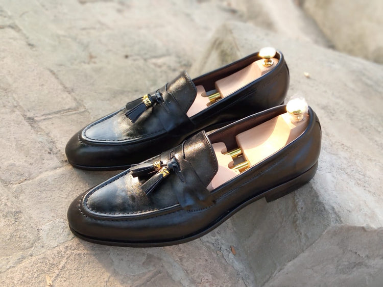 A pair of black tassel loafers with gold accents on a stone surface.